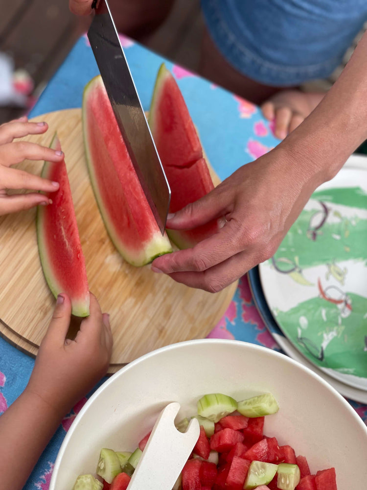 mums hands cutting Watermelon with children hands, bowl full of cucumber and water melon salad, green turtles printed plates