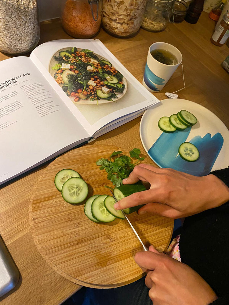 
                  
                    recipe open from book,cucumber sliced on wooden lid from bowl, herbal tea in bamboo cup and some slices of cucumber plate next to board , jars filled with ingredients , oats, lentils and jam ,in back ground
                  
                