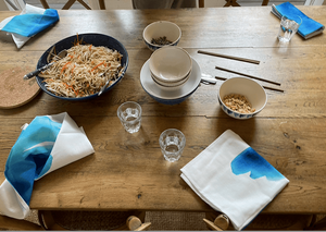 
                  
                    tablescape of noodles in salad bowl with small bowls, chop sticks , water cups, and  cotton napkins with blue wave prints.
                  
                